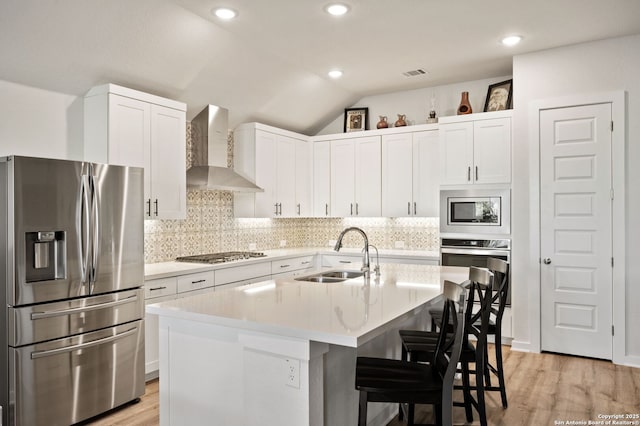 kitchen featuring stainless steel appliances, wall chimney range hood, light countertops, and a center island with sink