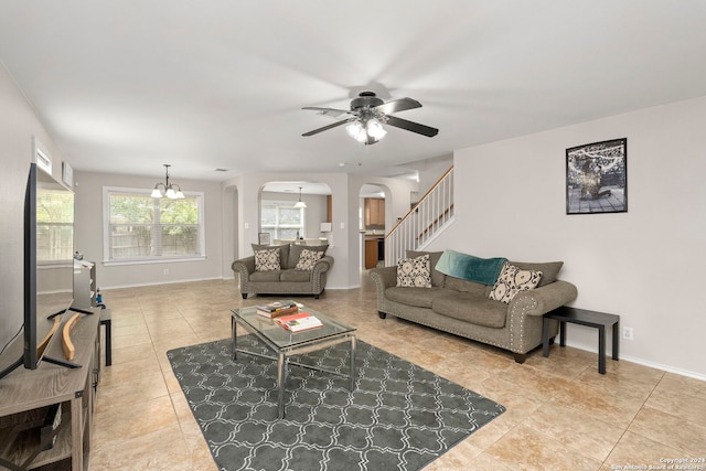 living room featuring arched walkways, light tile patterned floors, ceiling fan with notable chandelier, baseboards, and stairway