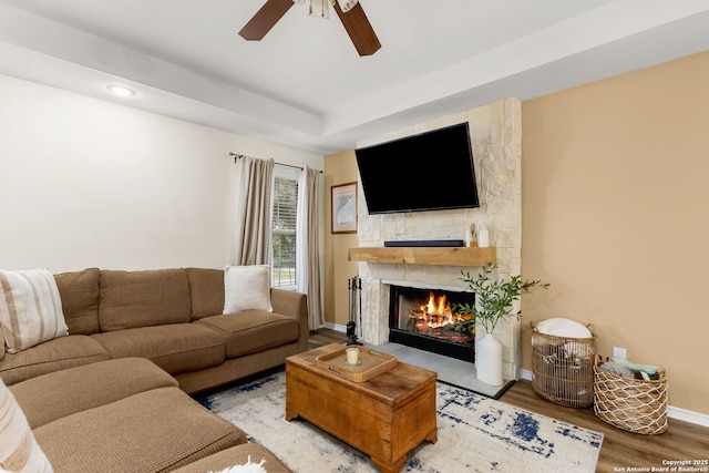 living room with a tray ceiling, a fireplace, a ceiling fan, wood finished floors, and baseboards