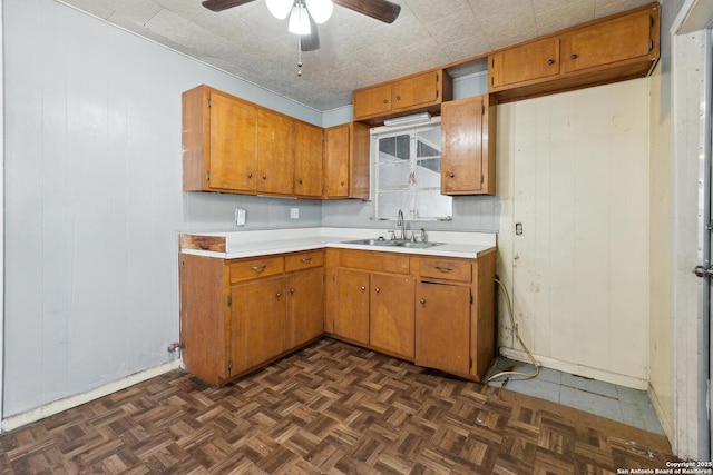 kitchen featuring wooden walls, a ceiling fan, brown cabinets, light countertops, and a sink