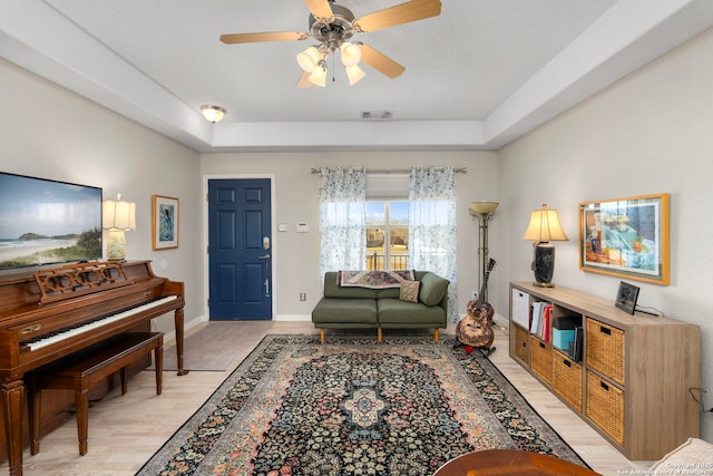 living room featuring ceiling fan, light wood-style flooring, visible vents, baseboards, and a tray ceiling