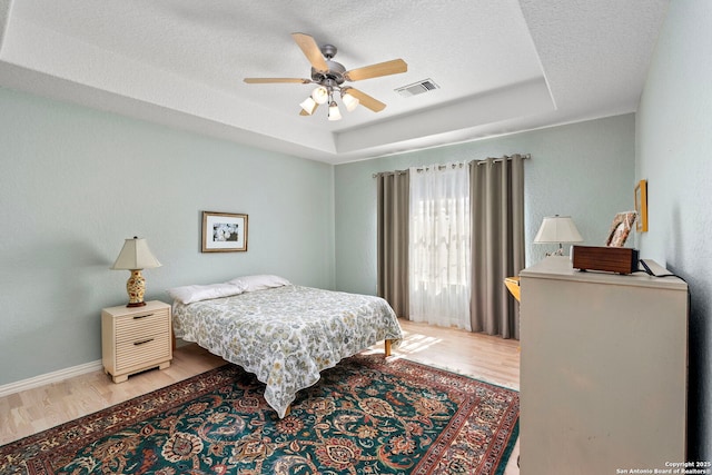 bedroom featuring a tray ceiling, visible vents, a textured ceiling, wood finished floors, and baseboards