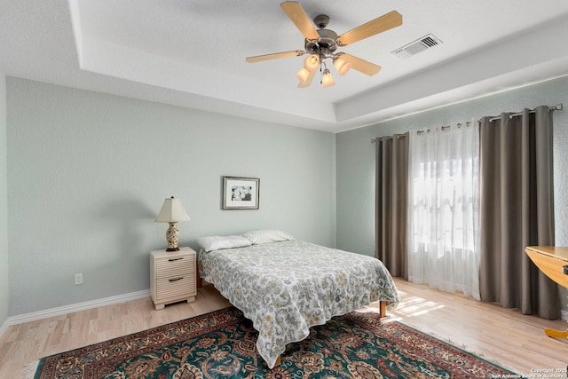 bedroom featuring a raised ceiling, visible vents, light wood-style flooring, a ceiling fan, and baseboards