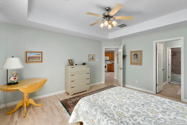 bedroom featuring light wood finished floors, a raised ceiling, visible vents, ensuite bathroom, and baseboards