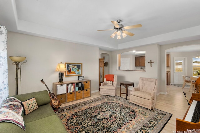 living area featuring arched walkways, a ceiling fan, baseboards, light wood finished floors, and a tray ceiling