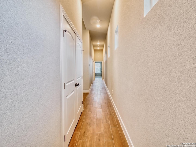 hall featuring a textured wall, light wood-style flooring, and baseboards