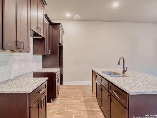 kitchen featuring decorative backsplash, an island with sink, dark brown cabinets, wood finish floors, and a sink