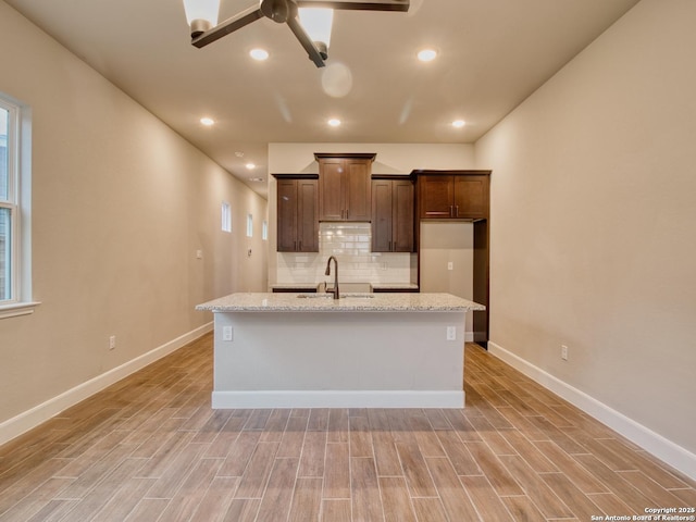 kitchen featuring dark brown cabinetry, a center island with sink, decorative backsplash, light wood-type flooring, and a sink