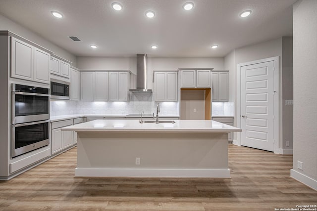 kitchen with light wood finished floors, double oven, a sink, built in microwave, and wall chimney exhaust hood