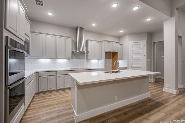 kitchen featuring double oven, black electric cooktop, a sink, wall chimney exhaust hood, and light wood finished floors