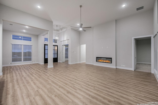 unfurnished living room with ceiling fan, visible vents, a towering ceiling, light wood-style floors, and a glass covered fireplace