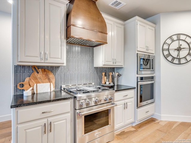 kitchen featuring light wood finished floors, stainless steel appliances, custom range hood, visible vents, and decorative backsplash