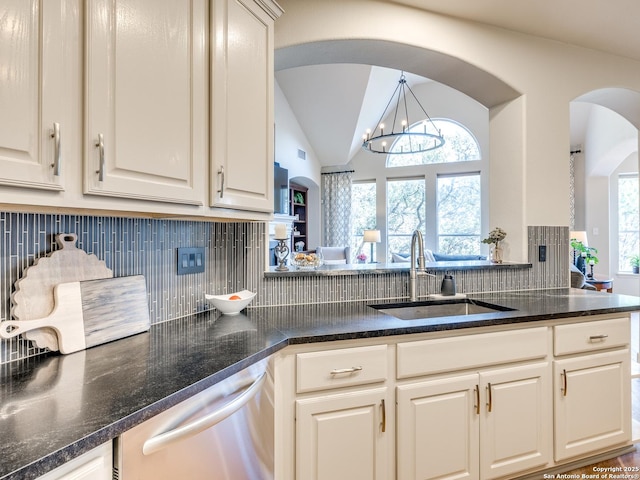 kitchen featuring a sink, stainless steel dishwasher, decorative backsplash, dark stone counters, and an inviting chandelier