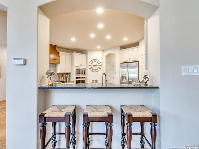 kitchen featuring arched walkways, a peninsula, premium range hood, appliances with stainless steel finishes, and a kitchen bar