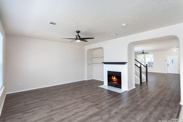 unfurnished living room with arched walkways, a fireplace with flush hearth, visible vents, a ceiling fan, and dark wood finished floors