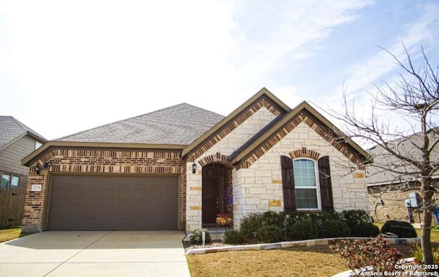 french country inspired facade with an attached garage, brick siding, concrete driveway, stone siding, and roof with shingles