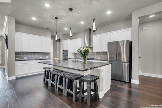 kitchen with wall chimney range hood, a center island with sink, appliances with stainless steel finishes, and dark wood-style flooring