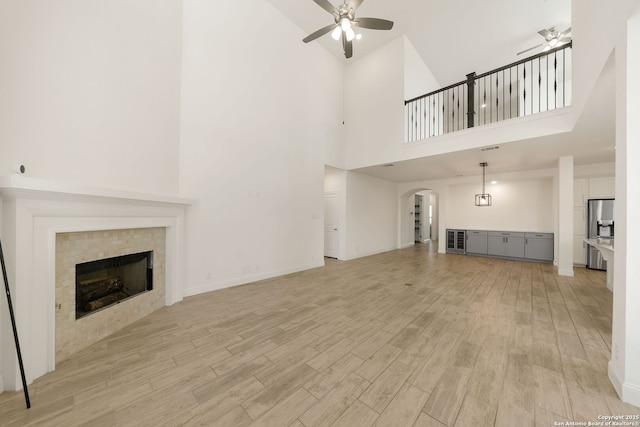 unfurnished living room featuring ceiling fan, arched walkways, baseboards, light wood-type flooring, and a tiled fireplace