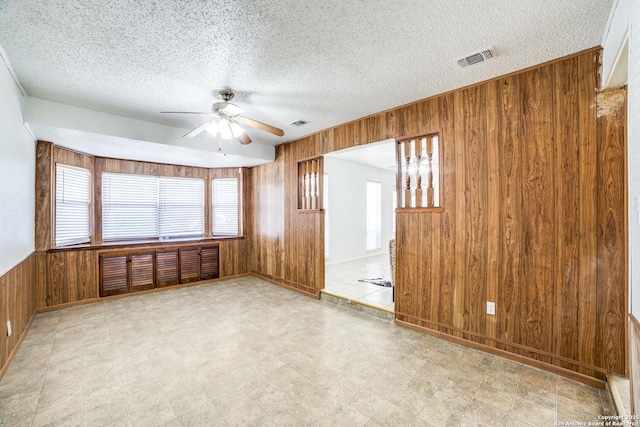 empty room with a textured ceiling, wooden walls, a ceiling fan, visible vents, and tile patterned floors