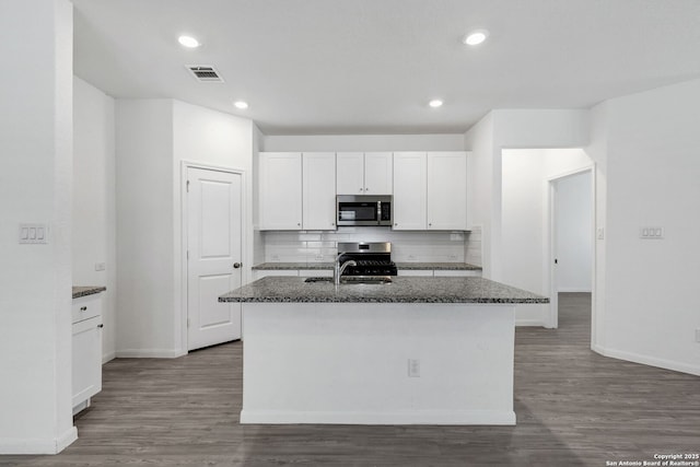 kitchen with stainless steel appliances, white cabinets, backsplash, dark stone countertops, and a center island with sink