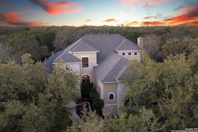 view of front of house with roof with shingles, a chimney, a wooded view, and stucco siding