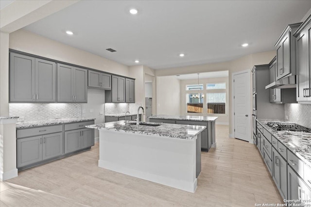 kitchen featuring backsplash, a sink, an island with sink, light stone countertops, and under cabinet range hood