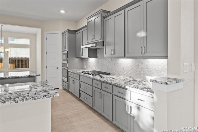 kitchen featuring light stone countertops, gray cabinetry, under cabinet range hood, appliances with stainless steel finishes, and decorative backsplash