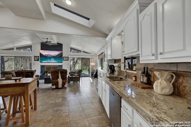 kitchen featuring stainless steel dishwasher, open floor plan, white cabinets, a sink, and vaulted ceiling with skylight