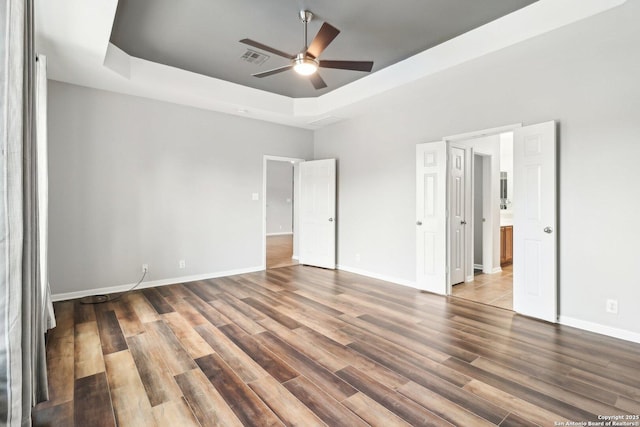 unfurnished bedroom featuring a tray ceiling, visible vents, ensuite bathroom, wood finished floors, and baseboards