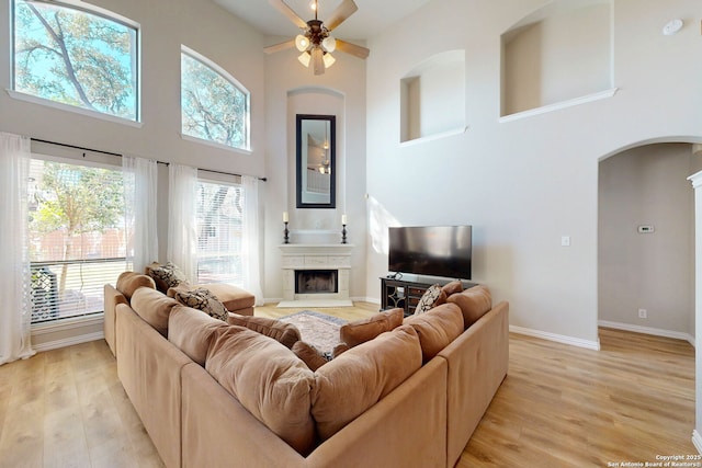 living room with arched walkways, a fireplace with raised hearth, baseboards, light wood-style flooring, and a high ceiling