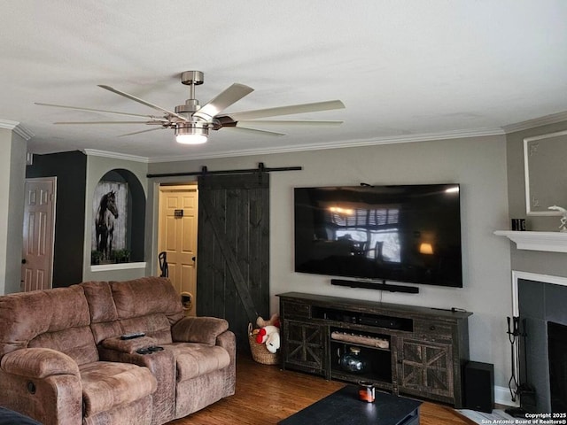 living room featuring a tiled fireplace, a barn door, a ceiling fan, ornamental molding, and wood finished floors