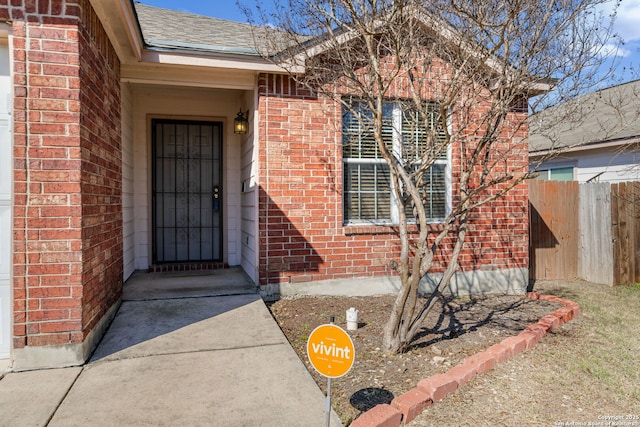 doorway to property with a shingled roof, fence, and brick siding