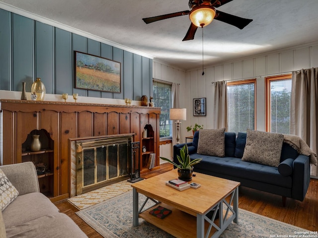 living room featuring a decorative wall, ornamental molding, a ceiling fan, a brick fireplace, and wood finished floors