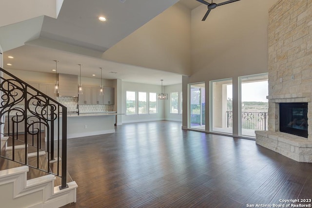 unfurnished living room featuring a towering ceiling, dark wood-style floors, stairway, a stone fireplace, and ceiling fan with notable chandelier