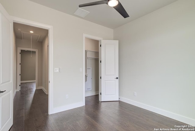 unfurnished bedroom featuring a walk in closet, dark wood-style flooring, attic access, ceiling fan, and baseboards