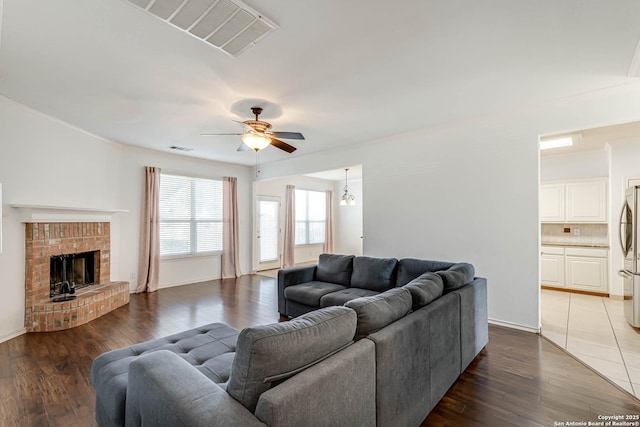 living area with a brick fireplace, ceiling fan, visible vents, and wood finished floors