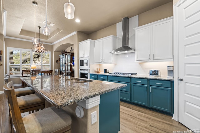 kitchen featuring arched walkways, blue cabinets, a sink, wall chimney range hood, and a tray ceiling