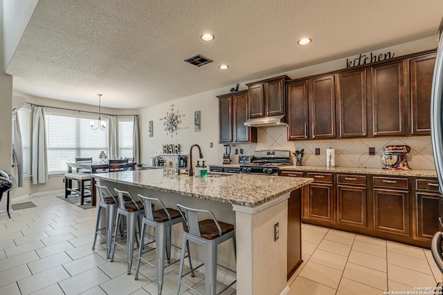 kitchen with a center island with sink, stainless steel gas stove, a sink, light stone countertops, and under cabinet range hood