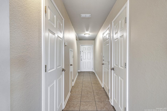 hall featuring light tile patterned floors, a textured wall, visible vents, and baseboards