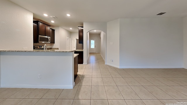 kitchen with arched walkways, light stone counters, recessed lighting, dark brown cabinets, and appliances with stainless steel finishes