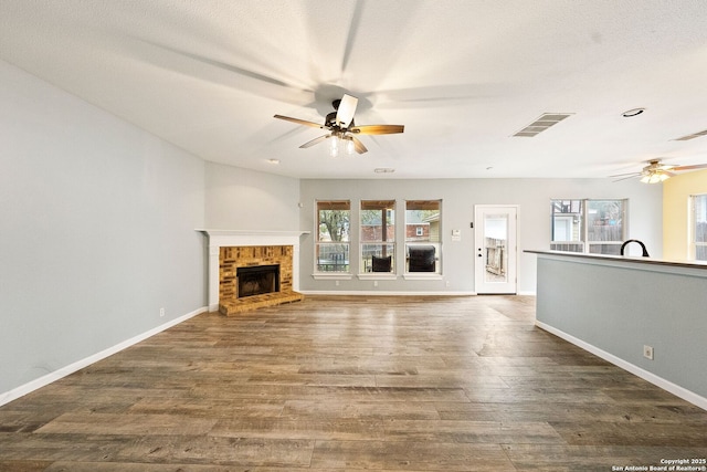 unfurnished living room with a brick fireplace, ceiling fan, visible vents, and wood finished floors