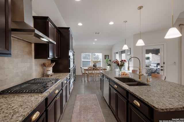 kitchen featuring a kitchen island with sink, a sink, appliances with stainless steel finishes, backsplash, and wall chimney exhaust hood