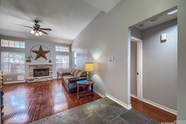 living room with ceiling fan, a stone fireplace, a textured ceiling, wood finished floors, and baseboards