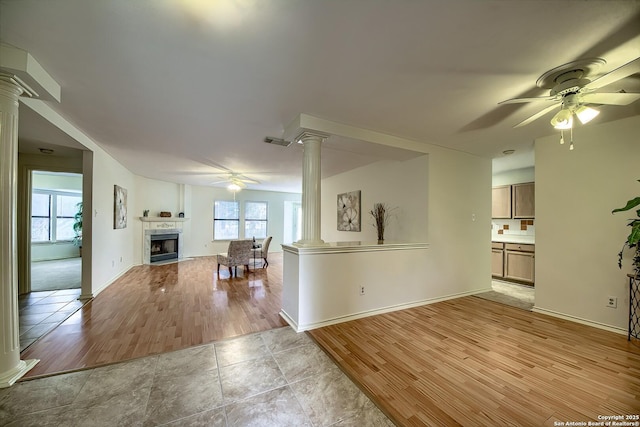 unfurnished living room with ceiling fan, a tile fireplace, visible vents, light wood finished floors, and ornate columns