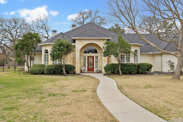 french country home featuring fence, stone siding, roof with shingles, a chimney, and a front yard