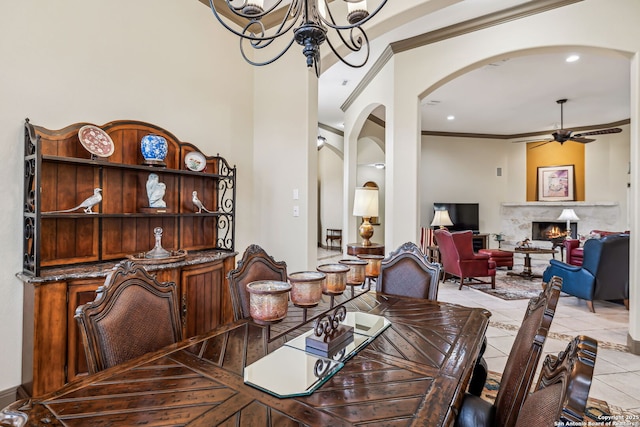 dining room with light tile patterned floors, arched walkways, a lit fireplace, crown molding, and ceiling fan with notable chandelier