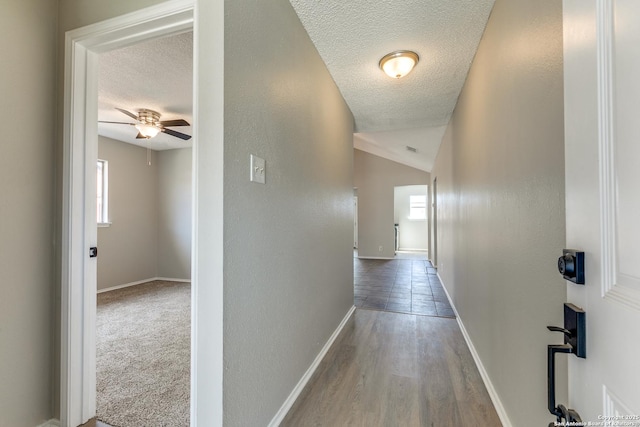 corridor featuring a textured ceiling, a textured wall, lofted ceiling, wood finished floors, and baseboards