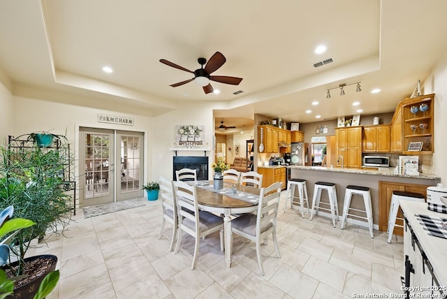 dining area with ceiling fan, a fireplace, visible vents, french doors, and a tray ceiling