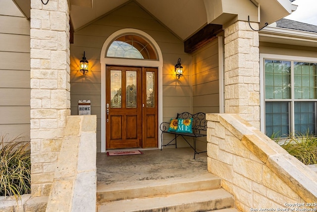 doorway to property featuring stone siding, a porch, and roof with shingles