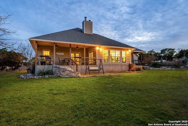 back of property with roof with shingles, a chimney, a ceiling fan, and a yard